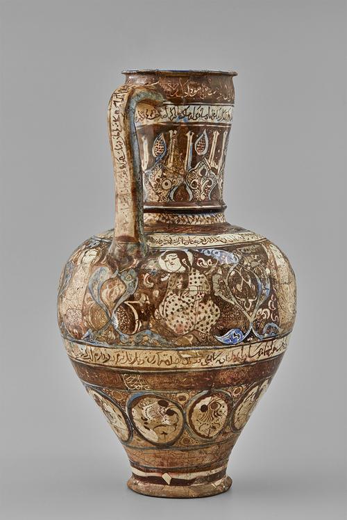 A ceramic jug with a slender handle, bulbous lower body, and rimmed opening. It is decorated with brown and blue against a white body, showing figures and floral medallions. There are five bands of inscriptions, with another inscription painted on the flat side of the handle.