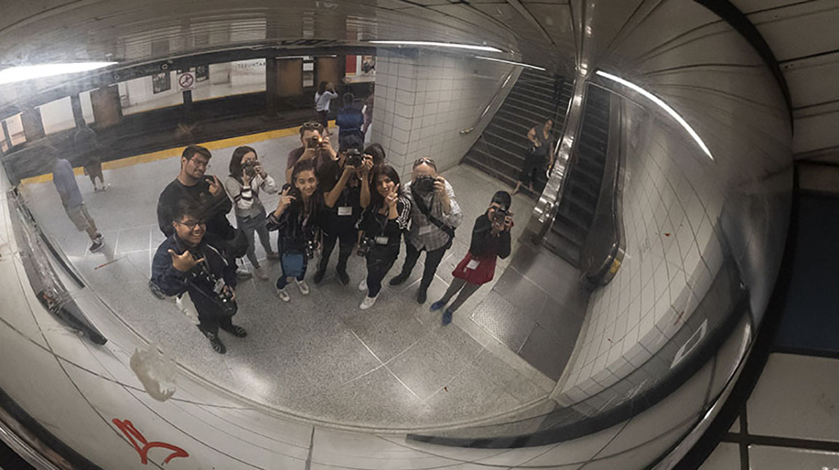 A line of student photographers standing on the subway platform looking up at a reflection of themselves in a mirror.