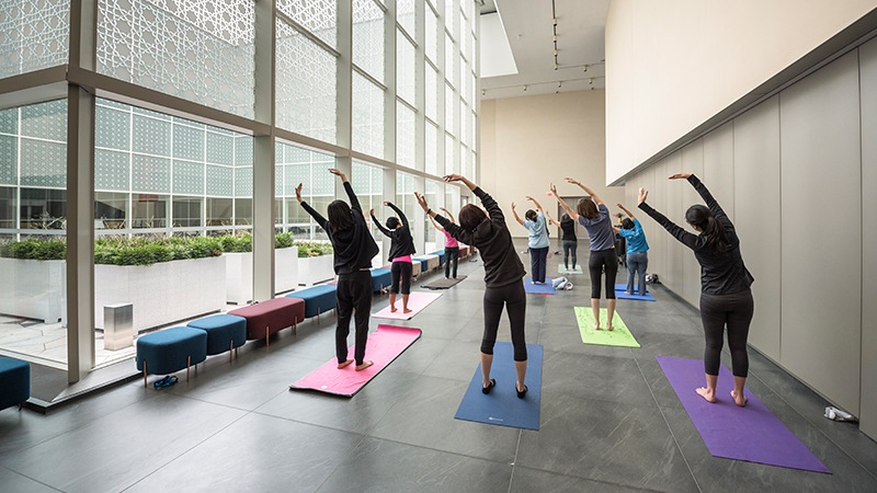 A group of women, facing away, lean to the left with arms raised, during a yoga class in the Museum's atrium.