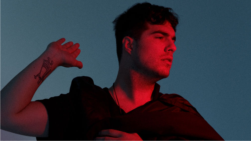 Jeremy Dutcher stands against a blue backdrop in an orange glow, wearing a brown cape and holding up his right hand.