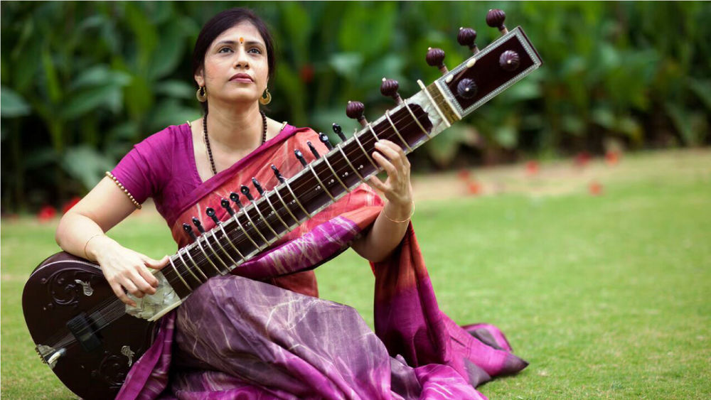Vidushi Anupama Bhagwat sits on grass outside, wearing a plum-coloured sari, playing the sitar.