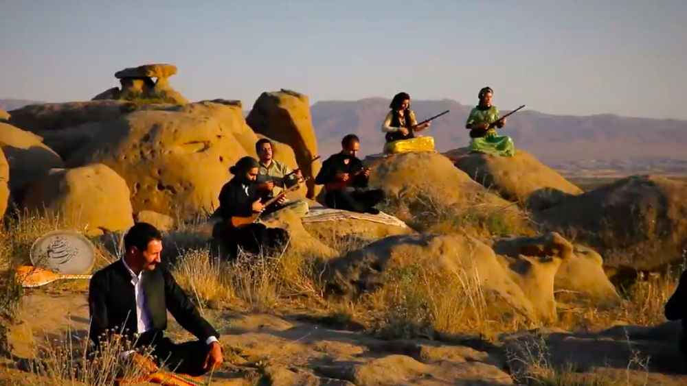 Four male musicians and two female musicians sit on rock formations, playing instruments in the setting sun.