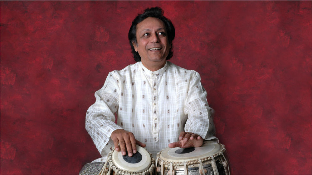 Swapan Chaudhari, wearing white, smiles and plays the tabla against a wine-coloured backdrop.