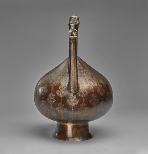 Brass kettle-shaped vessel mounted on a raised, circular base. View from the back of the large, semi-circular handle.