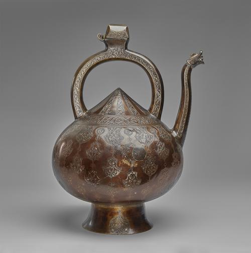 Brass kettle-shaped vessel with a pointed top. The long, upright spout has a roaring dragon head at the tip. The vessel is mounted on a raised, circular base. It has a large, semi-circular handle with a hinged opening at the top. It is decorated with engraved scrolling vine patterns.