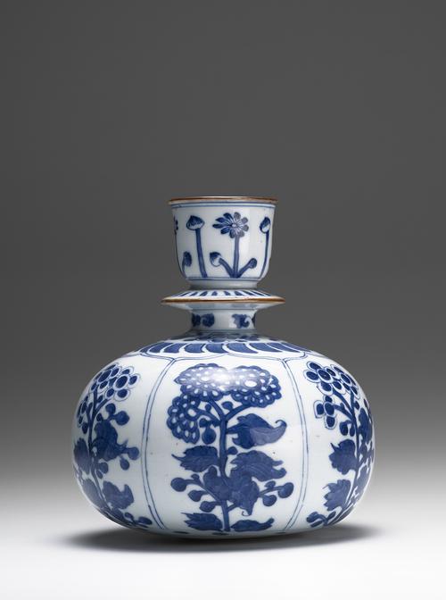White ceramic vessel with a large, rounded base and a thin neck with a wide, flat disc half-way to the mouth. There are blue flowers on the base, and five bands of flowers and rounded shapes decorate the neck. The mouth and the edge of the disc are brown.