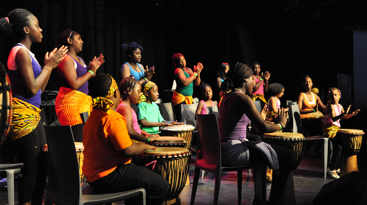 A group of children and teens, wearing colourful clothes, drum and clap on stage.