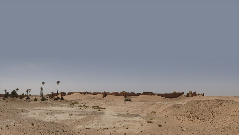 A desert landscape with sand dunes in the foreground and a line of ruined walls on the horizon.