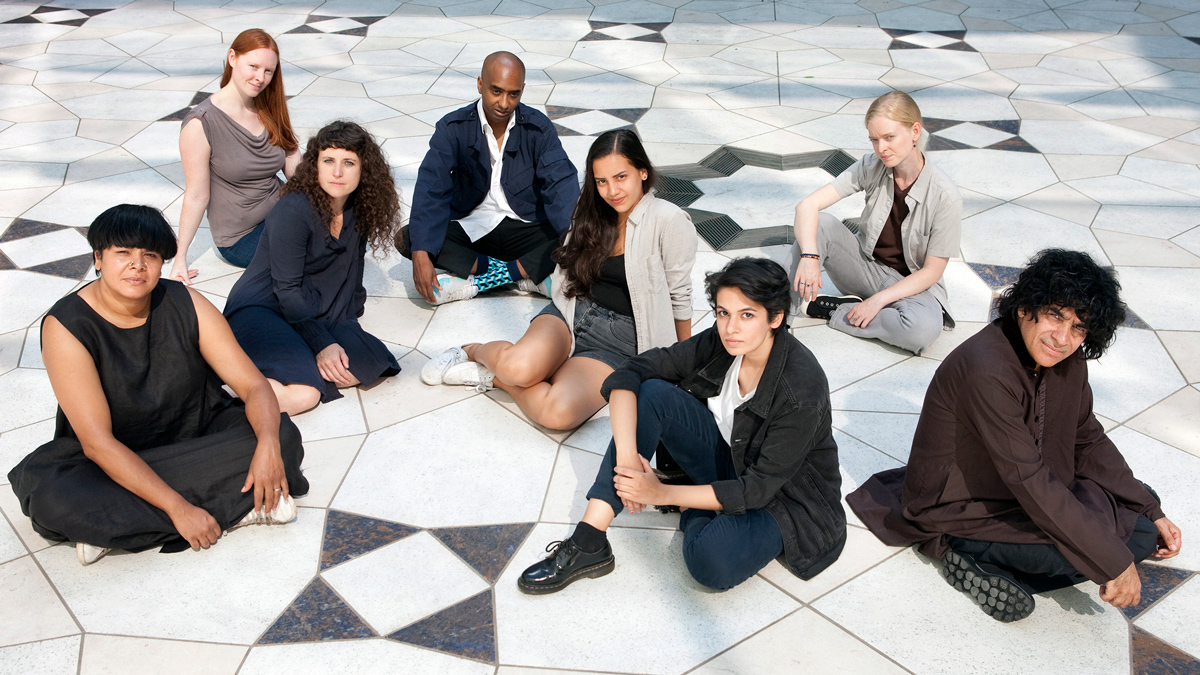 A group of eight young women and men pose seated on the tiled, geometric-patterned floor of the Museum’s courtyard during the day. 