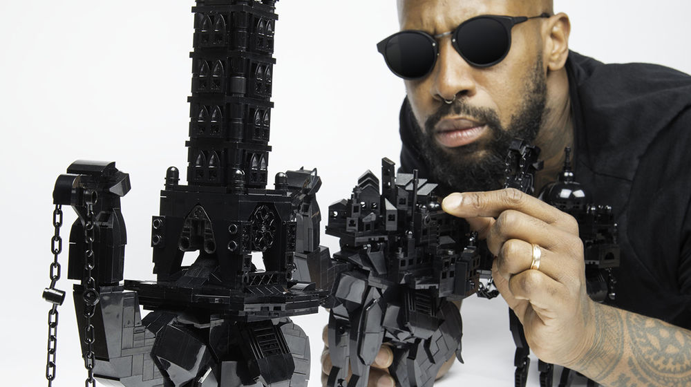 A man in sunglasses crouches down to touch a camel sculpture made of shiny black Lego pieces.