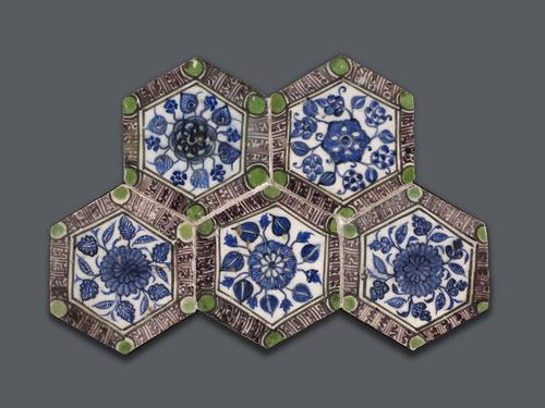 A set of five hexagonal tiles, painted white and decorated. Each features a large central floral rosette with various types of leaves extending outward, all in blue. The borders feature a band of purple script with large bright green circles at each point of the hexagon.