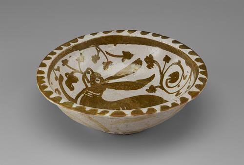 View of a circular bowl, decorated in softly metallic gold paint over an off-white base. The flattened rim is decorated in a scalloped pattern, with a large, floppy-eared hare surrounded by flowers in the centre. The outside is decorated with soft daubs of paint.