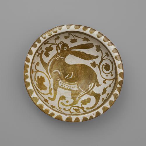 Top view of a circular bowl, decorated in softly metallic gold paint over an off-white base. The rim is decorated with a scalloped pattern, while in the centre is a large, floppy-eared hare surrounded by flowers.