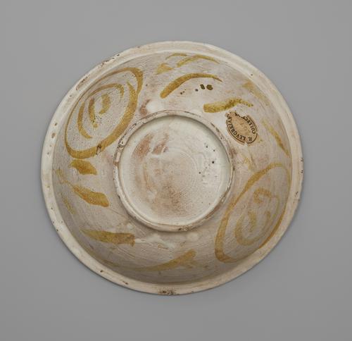 Bottom view of a circular bowl, showing a slightly raised base and a flattened rim. The bowl is painted off-white, with circular lines and daubs of soft golden paint. There is an oval paper sticker on one edge that reads “H. Kevorkian Collection” with “2672” written on it in pencil.