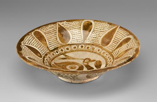 View of a cream bowl, decorated with gold paint. The base is small and raised. The interior depicts a bird holding a leaf, enclosed by a narrow band filled with circles. Large gold teardrops alternate direction (pointed in or out) around this band, with the spaces filled with v-shaped marks.