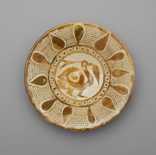 View of the interior of a cream bowl, decorated with gold paint on a cream background. In the centre is a bird holding a leaf, enclosed by a narrow band filled with circles. Around this band is a pattern of large teardrops, alternating direction, with v-shaped marks filling the spaces. 