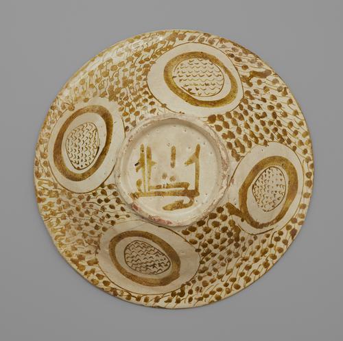 Bottom view of a circular bowl, showing the raised base, which is smaller than the rim’s diameter. The base is painted cream, with the Arabic word “barakah” painted on it. The exterior is decorated with four roundels, filled with v-shaped marks, set against a ground of irregular dots.