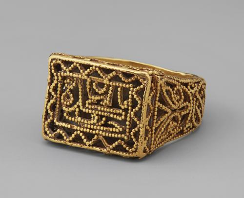 Golden rectangular ring laying on its side, top view of the rectangle shaped top and the band, the ring is granulated and uses filigree for decoration.