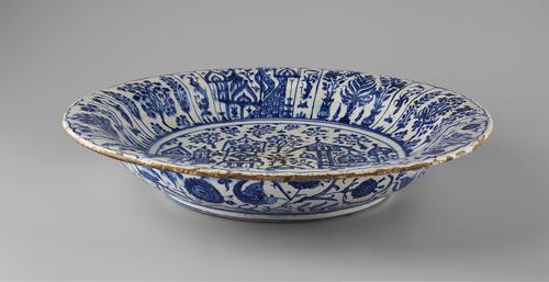 Side view of a dish with sloping rim, white base decorated with cobalt blue outlined in blue-black, the center of the dish depicts a cityscape with foliage, the rim has panels of trees and foliage.