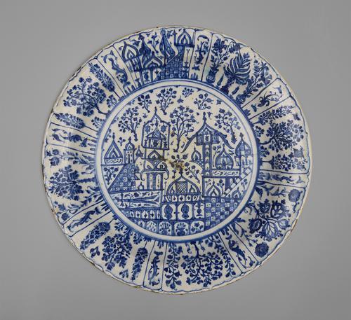 Top view of a dish with sloping rim, white base decorated with cobalt blue outlined in blue-black, the center of the dish depicts a cityscape with foliage, the rim has panels of trees and foliage.