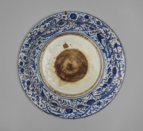Bottom view of dish with sloping rim, white base decorated with cobalt blue outlined in blue-black, the rim has panels of trees and foliage. The foot of the dish remains white with a brown round centre.