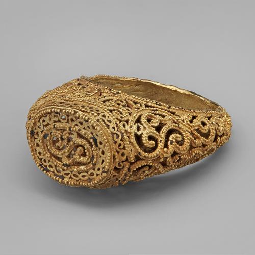 Golden ring laying on its side, view of the top oval bezel and flat top, featuring filigree and granulation, with arabesques widening towards the bottom of the band.