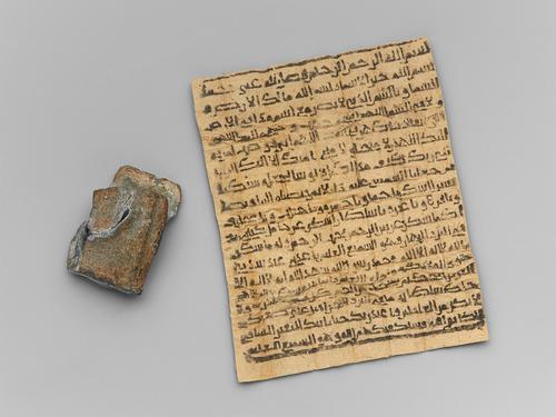 Square lead case, tarnished brown, beside a piece of tan paper that was once folded many times to fit into the case. Written in black on the paper are lines of Qur’anic verses that fill from edge to edge of the paper.