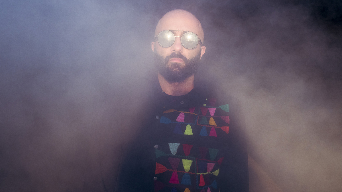 Narcy, wearing sunglasses and a multi-colored embroidered jacket, appears from within a cloud of smoke.