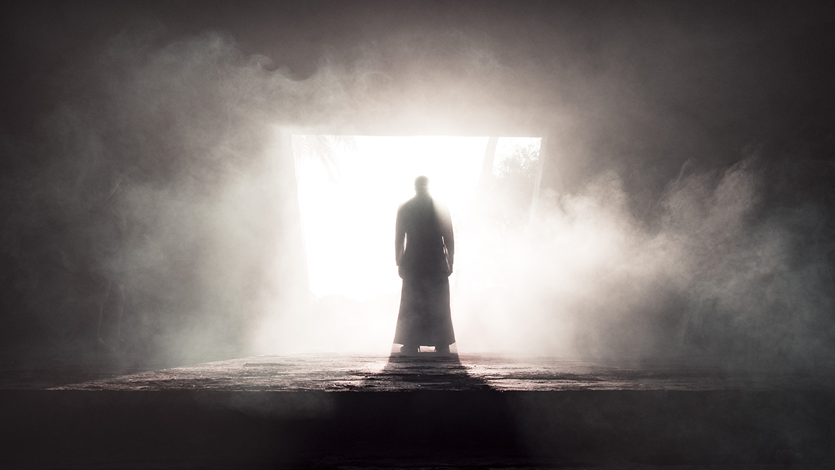 A silhouette of Narcy standing in a bright beam of light streaming in from an opening in the wall of a dark, smoky room.