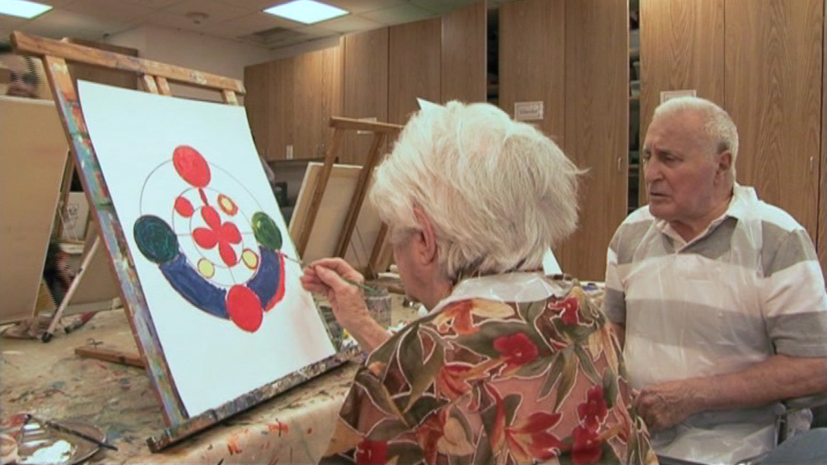 Inside a studio, an elderly woman works on a circular painting, as an elderly man looks on.