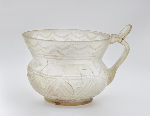 Side of a clear glass vessel with a squat globular body with a high, flaring neck nearly as tall as its body. A flat handle attached at the center of the body has thumb piece  at the rim of the neck. Decorated with zigzag lines and rows of crescents around the neck.