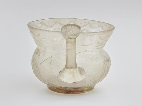 Clear glass vessel with a squat globular body with a high, flaring neck nearly as tall as its body.  Direct view of the flat handle and thumb piece visible at the rim of the neck. Decorated with zigzag lines and rows of crescents around the neck
