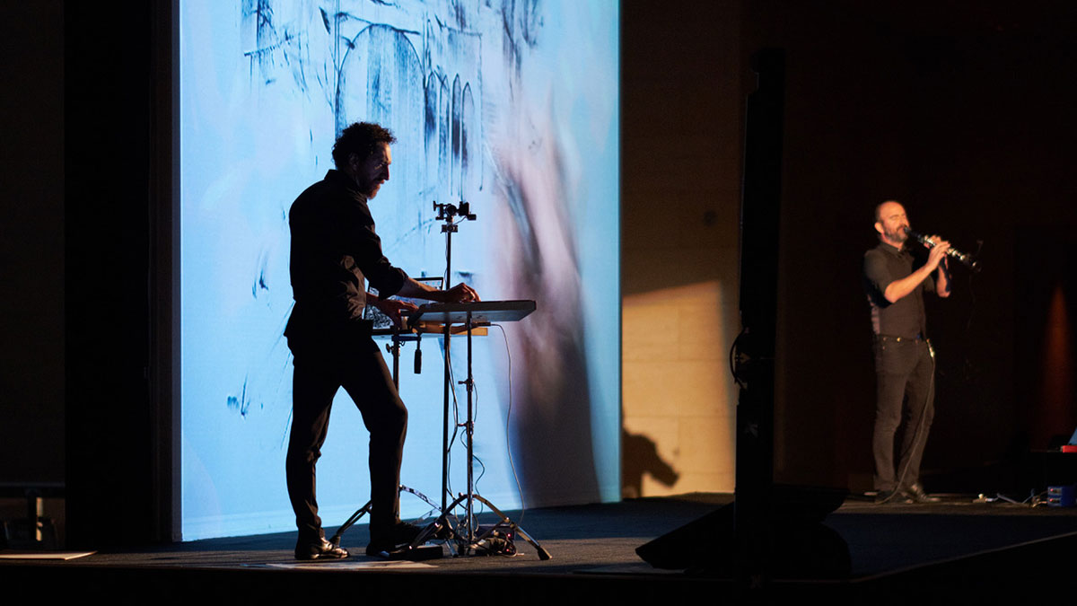 On stage, an artist draws at a table as the image appears on a large screen, while a clarinetist plays on the side.