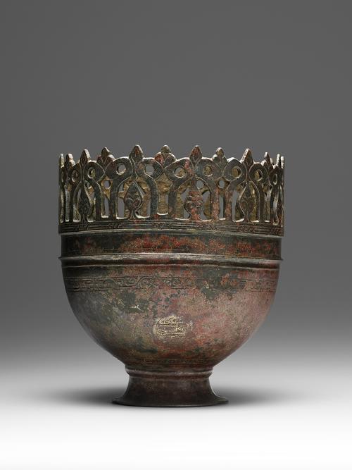 Metal bowl with tall foot, around the top a perforated crown decoratiion which features curling leaves and palmettes surmounted by cuspidate (pointed) leaves. On its body, an engraved band decorated with plant sprays appears above three medallions bearing plant motifs.