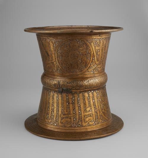 Brass; engraved cylindrical tray stand with silver and gold. A bold Arabic inscription decorates the upper and lower parts of the stand, narrower in the middle at the rib also featuring an Arabic inscription. Large roundel shown on upper part of the stand.