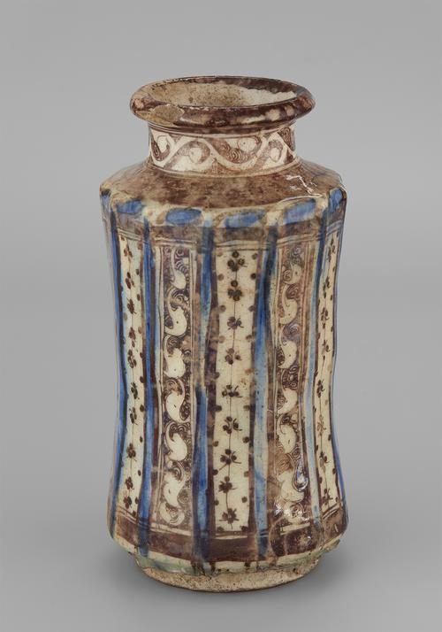 Brown and blue lustre on a cream coloured ceramic vase. Its concave body is divided by thirteen ridges painted in blue. The areas between contain panels of brown and white, either white arabesques on a brown ground or clusters of brown dots on a white ground. A band of brown arabesque circles the neck of the vessel