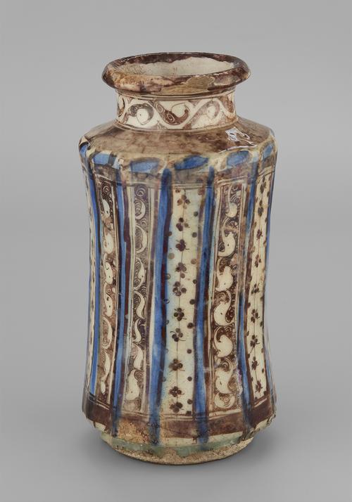 Brown and blue lustre on a cream coloured ceramic vase. Its concave body is divided by thirteen ridges painted in blue. The areas between contain panels of brown and white, either white arabesques on a brown ground or clusters of brown dots on a white ground. A band of brown arabesque circles the neck of the vessel.