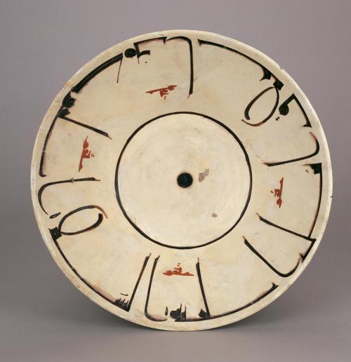 White dish of a truncated conical form, painted in manganese black with a monumental kufic inscription with secondary inscriptions in red slip, centred on a black spot in a single line roundel.
