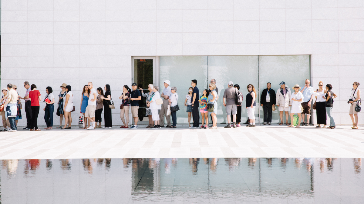 Forty people stand in line outside the Aga Khan Museum on a sunny summer day, with their reflections visible in a pool in the foreground.