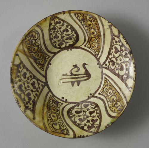 Bowl with a small calligraphic bird in the centre is painted in dark brown on a white slip. On the sides, variously shaped panels are filled with dots, peacock’s eyes and hearts.