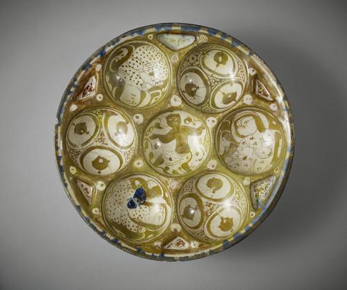Low dish containing seven hollows circles, six grouped around a central one. It is painted in brown lustre on a white ground. Four four of the six holows have figures decorating them. The rim of the dish is spotted brown and blue.