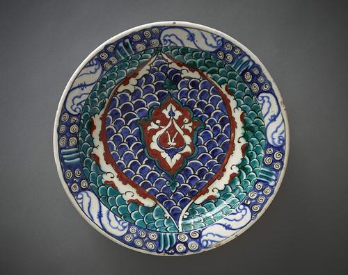 Ceramic dish with a slightly inverted rim, decorated in the centre with two long stemmed arabesque leaves forming an ogival motif and arched medallion with scalloped edges. The background decorated with a fish-scale motif in blue, green and red. The rim has scrollwork and cloud-shaped motifs.   