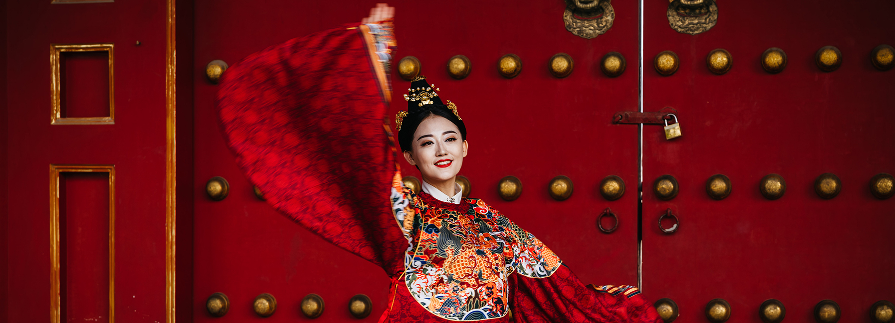 A young woman in a predominantly red traditional Chinese robe dances in front of a red and gold wall.