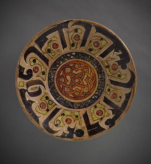 Dish with sloping rim, pinkish-brown ground with black, brick-red, yellow and white colours. Decorated with a bold calligraphic design interspersed with circular scroll motifs with larger dots. In the centre a design of interlacing strap-work with dotted interstices in red-brick and yellow colour.