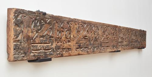 Side of wooden beam carved with kufic calligraphy and floral decoration, mounted on wall.