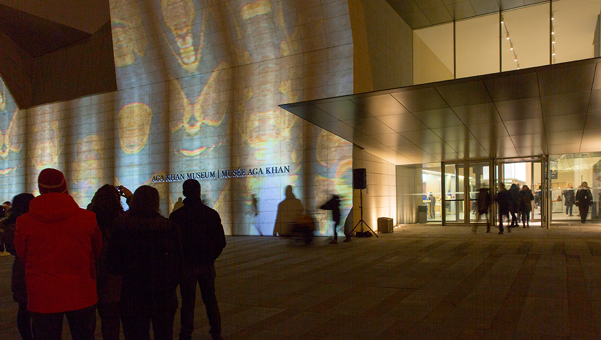 People outside at night looking at blue, green, and gold light projection illuminating the west facade of the Aga Khan Museum in Toronto. 