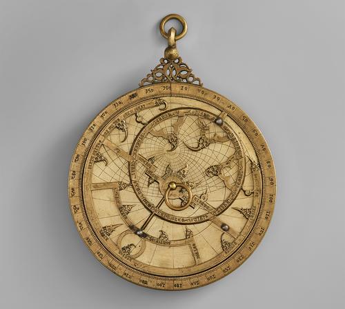 Front of the astrolabe, gold circular object, multiple plates stacked on top of each other with a pin through the middle and with a loop at the top.
