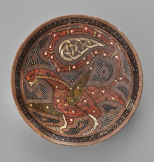 Bowl of shallow form, decorated in manganese-brown, tomato-red, white and olive-green slip on a purple ground with the figure of a fabulous bird with palmette wing and arabesque tail, a calligraphic band around the rim.