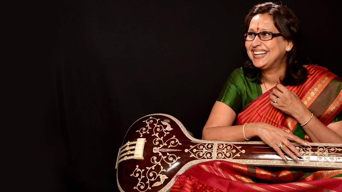 Arati Ankalikar-Tikekar sits smiling while holding a sitar and wearing a green and red outfit and glasses against a black background.