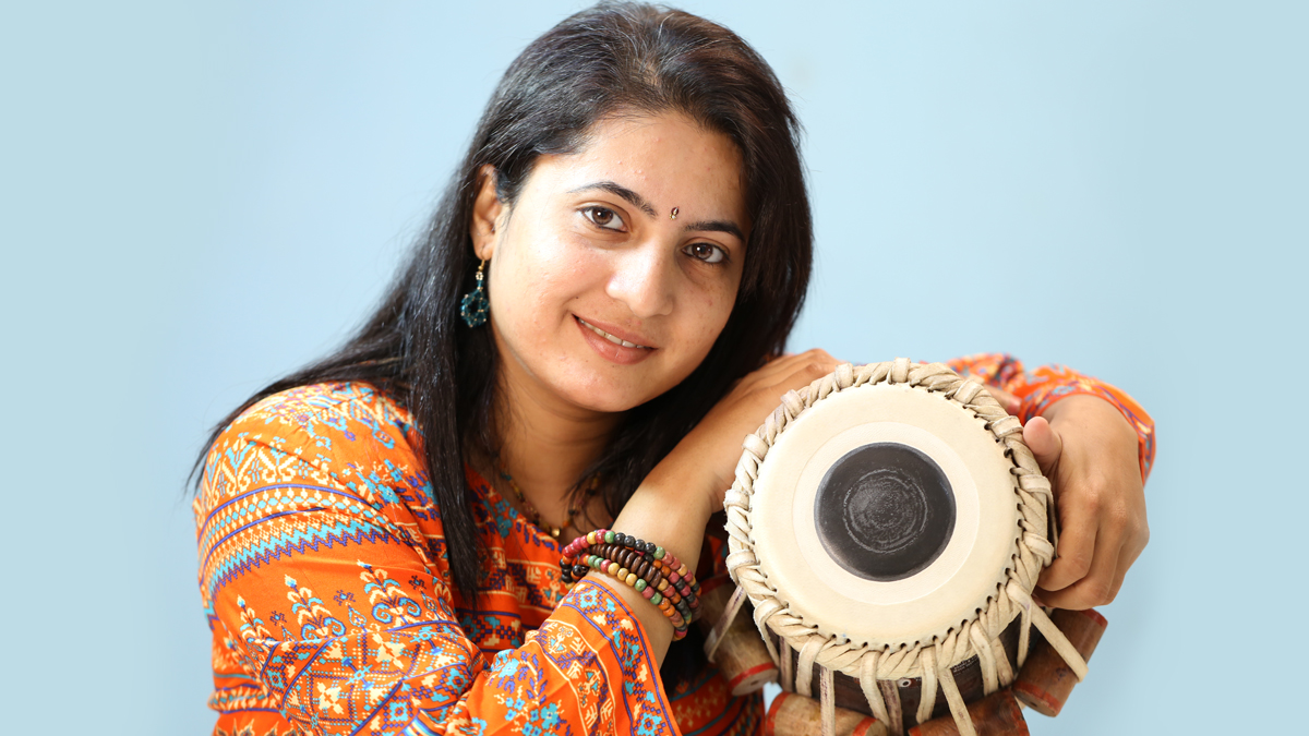 Mukta Raste, wearing an orange top and multicolored bracelet holds a tabla drum against a blue backdrop.